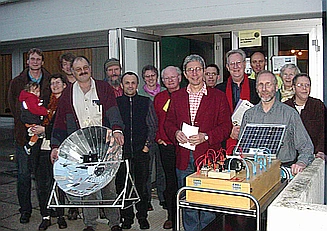 Abgebildet ist eine Gruppe Personen während einer Mitgliederversammlung in Regensburg. Ein Solarkocher und der visualisierte Aufbau einer PV-Anlage bilden den Vordergrund.