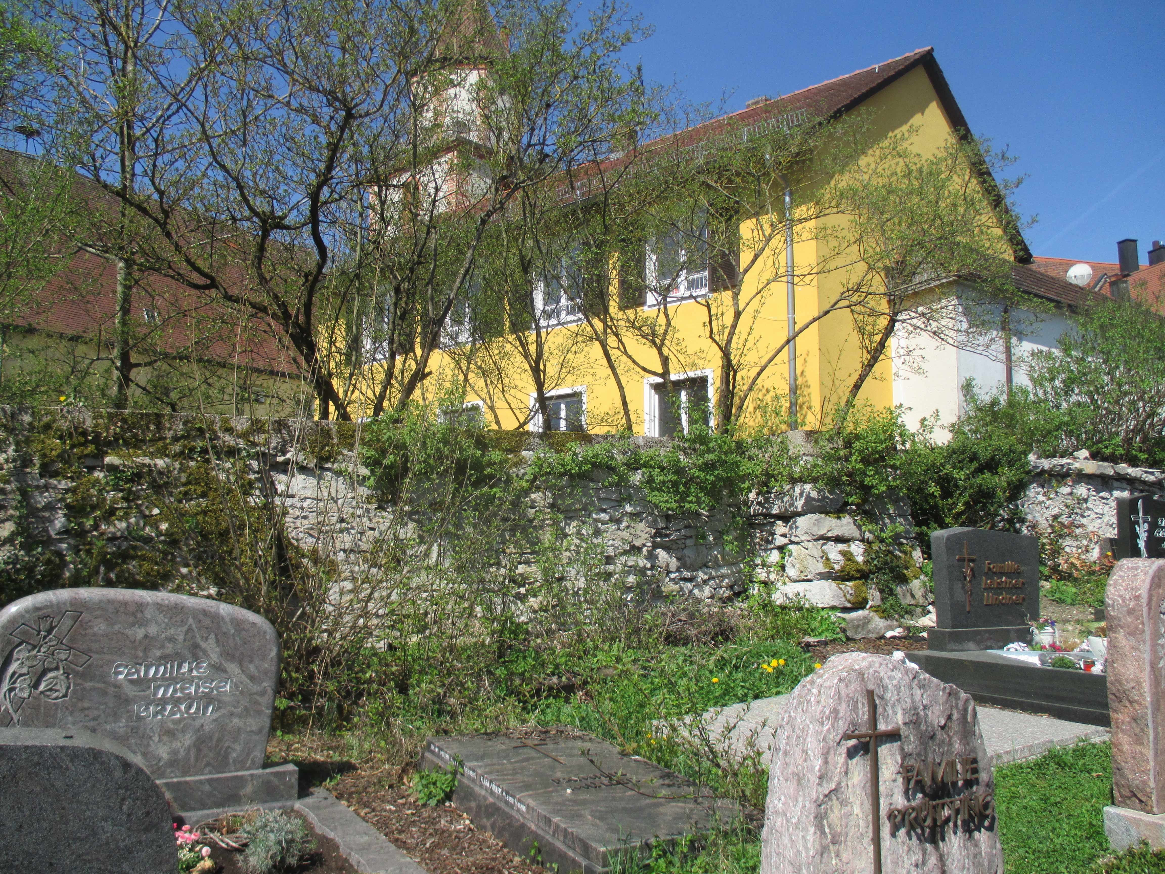 Eine bewachsene, alte Bruchsteinmauer begrenzt eine Friedhof hin zur Kirche.