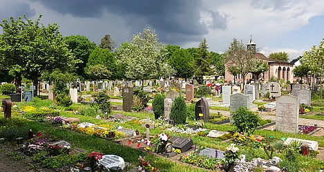 Blick auf einen Friedhof mit vielen Grabsteinen und Bepflanzung. Im Hintergrund eine kleine Kapelle und am Himmel zieht ein Gewitter auf.