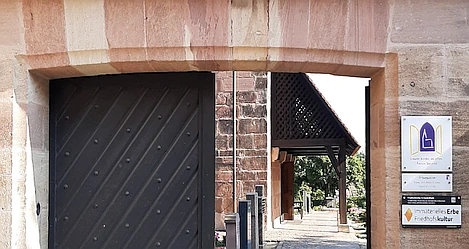 Das halb geöffnete große Eingangstor aus Holz zum Oasenfriedhof in St. Jobst gibt den Blick in den Friedhof frei. Zu sehen sind hier einige Grabsteine. 