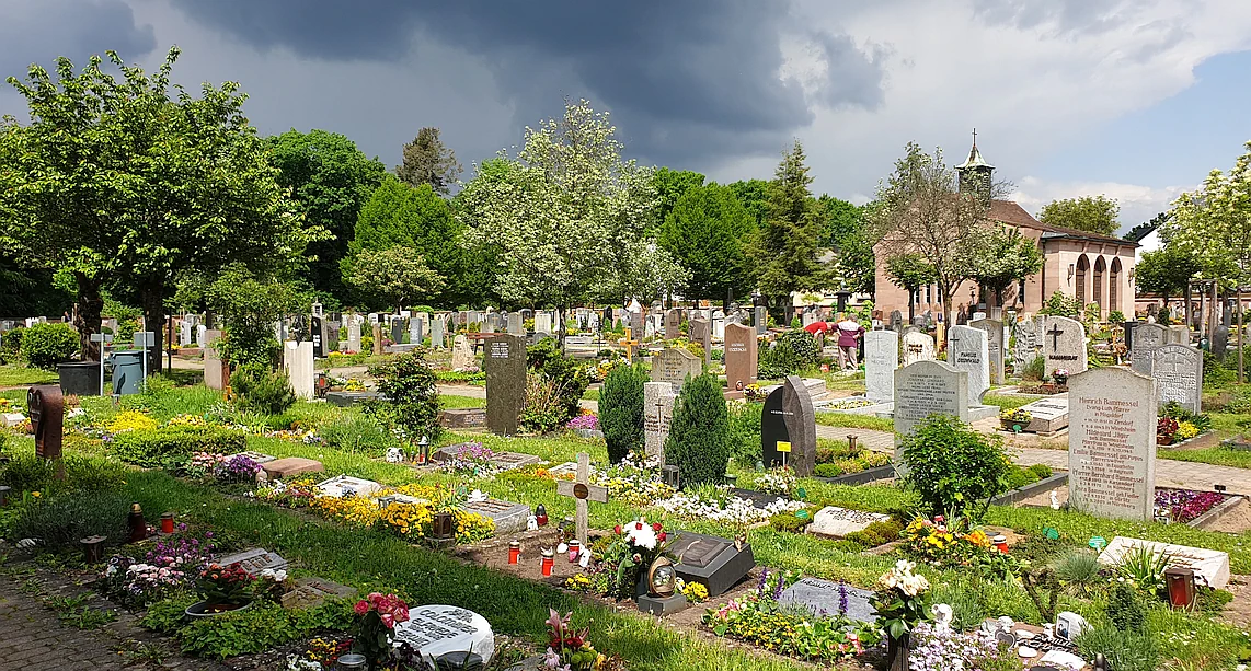 Blick auf einen Friedhof mit vielen Grabsteinen und Bepflanzung. Im Hintergrund eine kleine Kapelle und am Himmel zieht ein Gewitter auf.