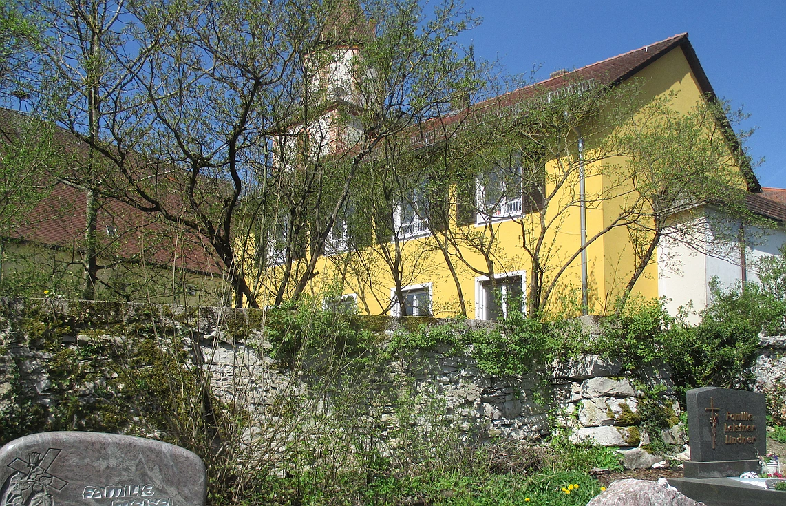 Eine bewachsene, alte Bruchsteinmauer begrenzt einen Friedhof hin zur Kirche.